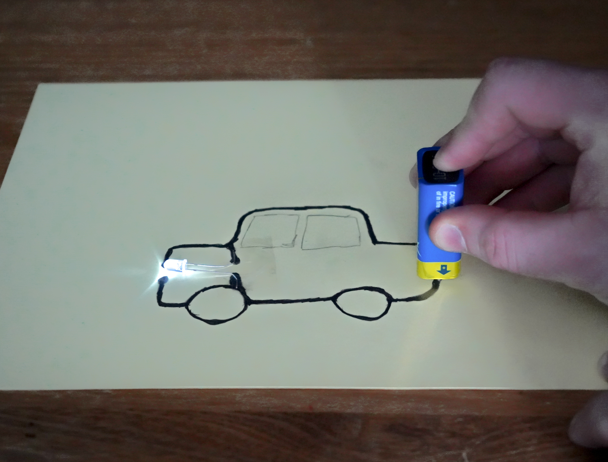 A car is drawn with two thick dark pencil lines on a piece of paper. A 9-volt battery is held by a hand at the back of the car, touching the drawing. At the front of the car, the two long metal legs of a white LED are taped to the paper. The LED is lit, like the headlights of a car.