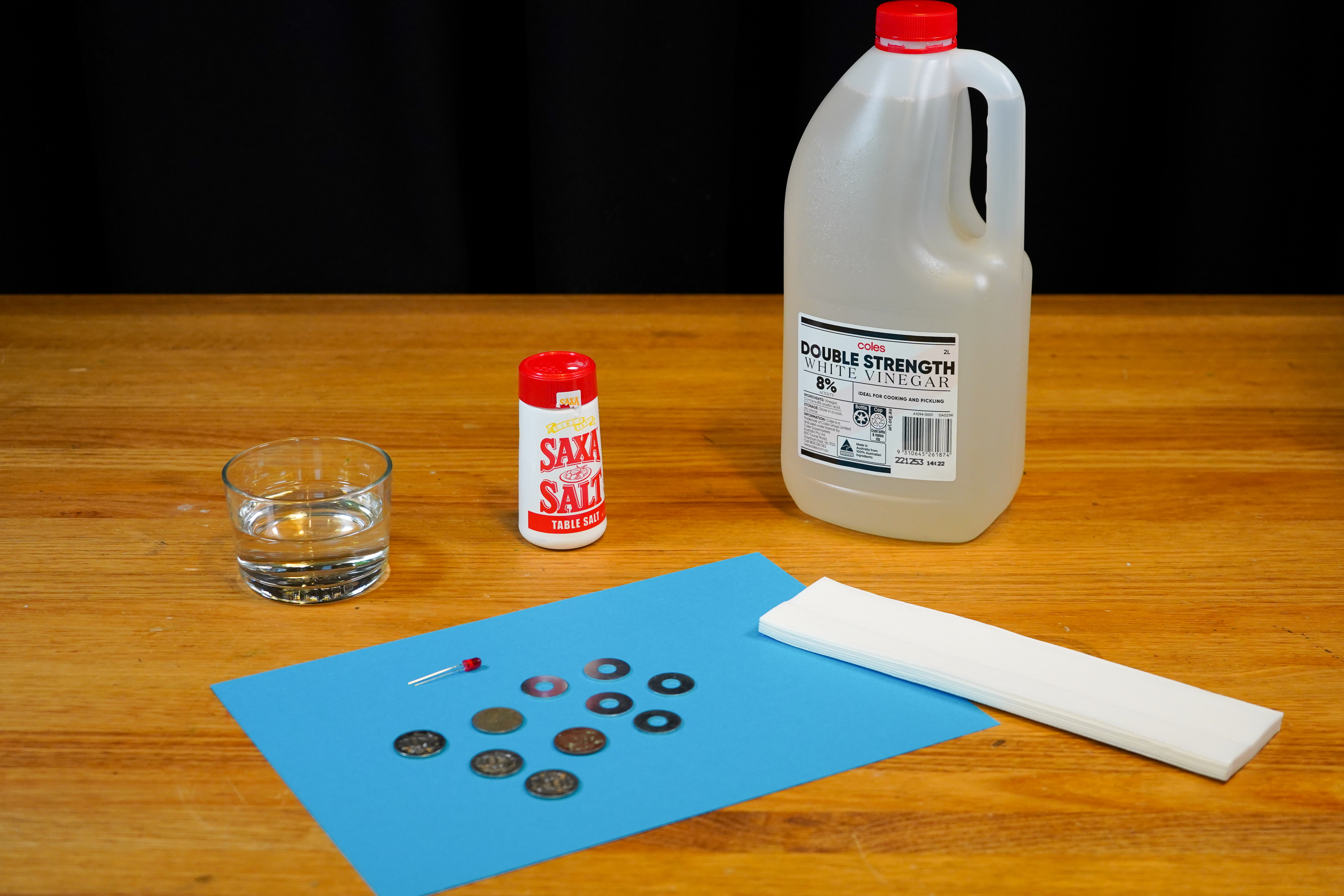 The materials for this experiment on a wooden table. Materials include a bottle of vinegar, table salt, a cup of water, a red light emitting diode, five washers, five ten cent coins, and paper towel.