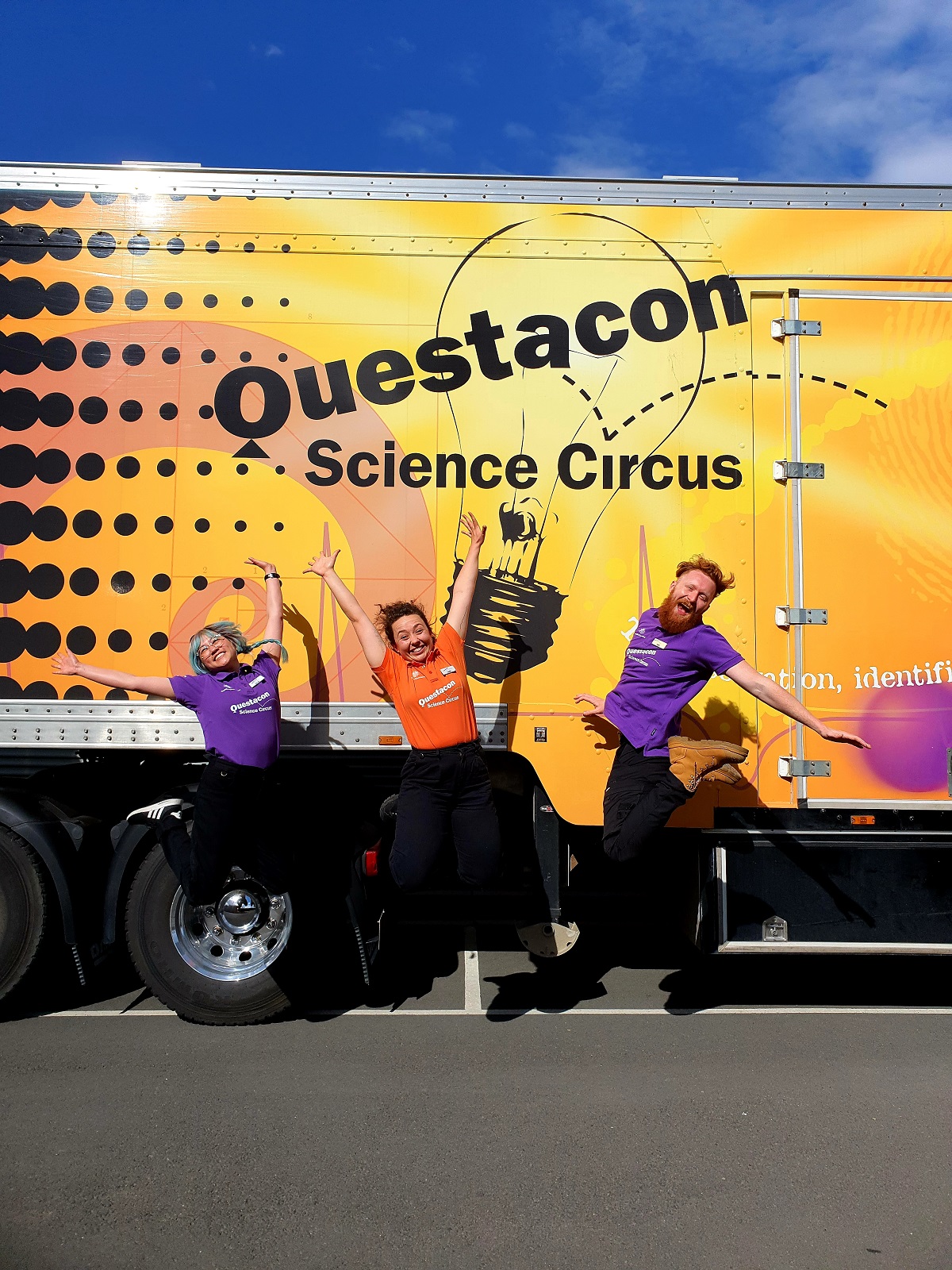 Three young people with excited expressions jumping with arms stretched wide. The questacon Science Circus bus is in the background