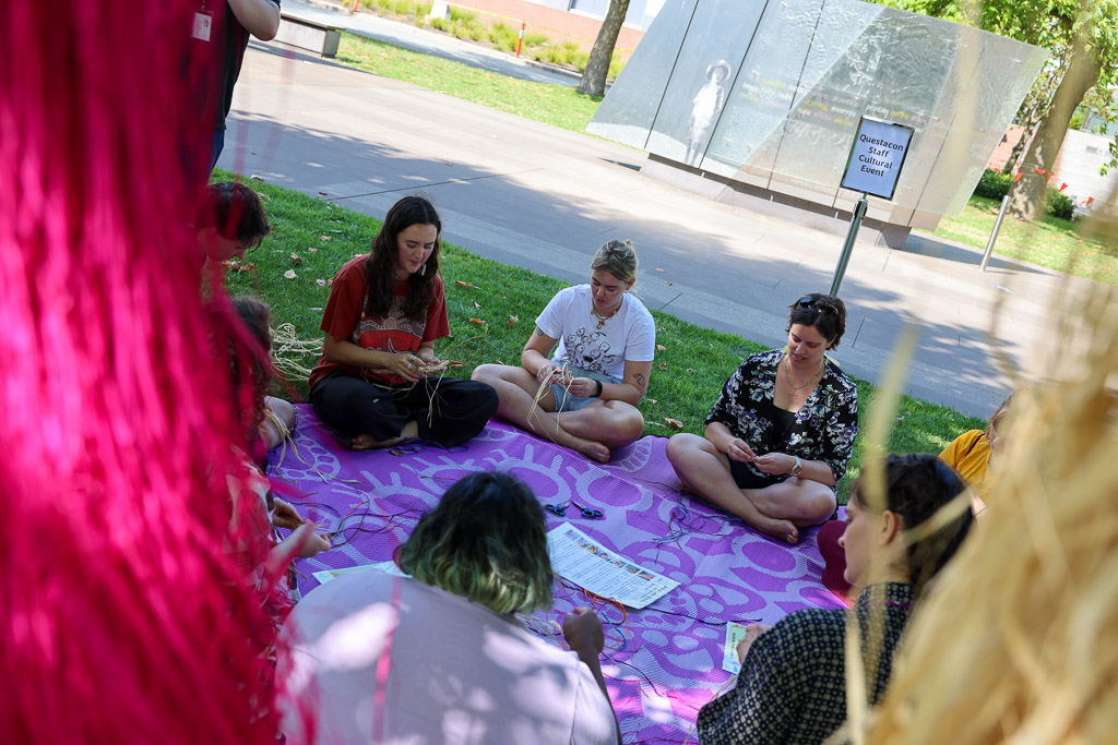 A group of women seated on a purple mat are weaving threads