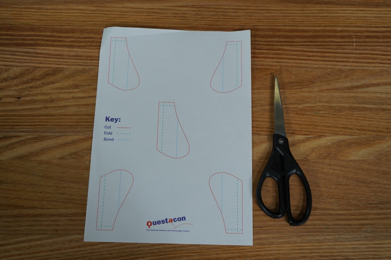 A top-down view of an A4 piece of paper and a pair of scissors on a table. A set of wing-shaped templates are printed on the paper, with lines showing where to cut, fold, and bend the paper. 