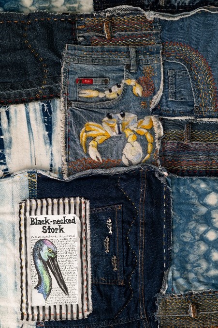 A close-up image of a denim patchwork quilt created by artist Jasmine Jan. The quilt features an embroidered crab, and an illustrated picture of a bird.