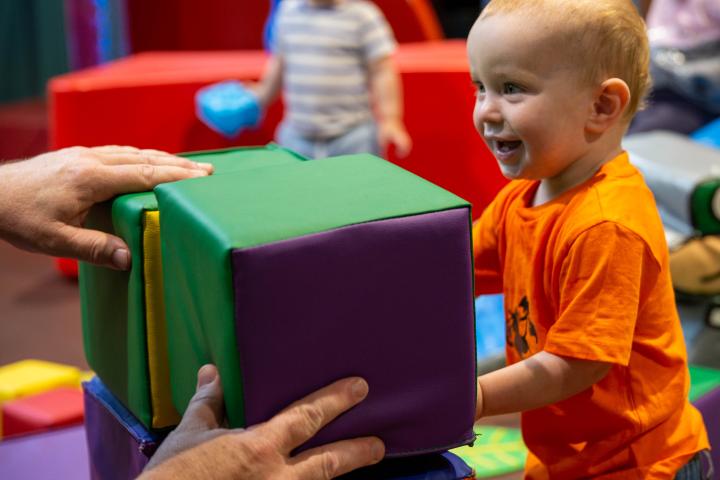 A young child in an orange t-shirt is playing with soft, coloured square blocks with their caregiver.