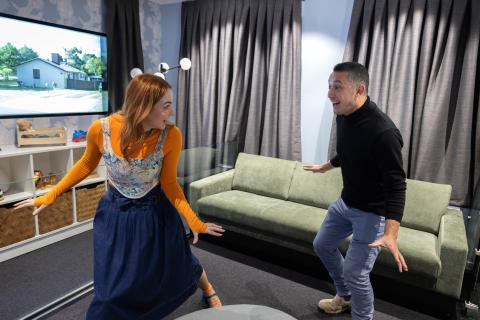 A woman and a man are in the earthquake house living room with their arms apart as they brace for the simulated shake of an earthquake.