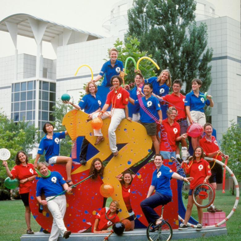 18 young adults dressed in blue and red shirts sit and stand around a bright statue of a cyclist outside Questacon.