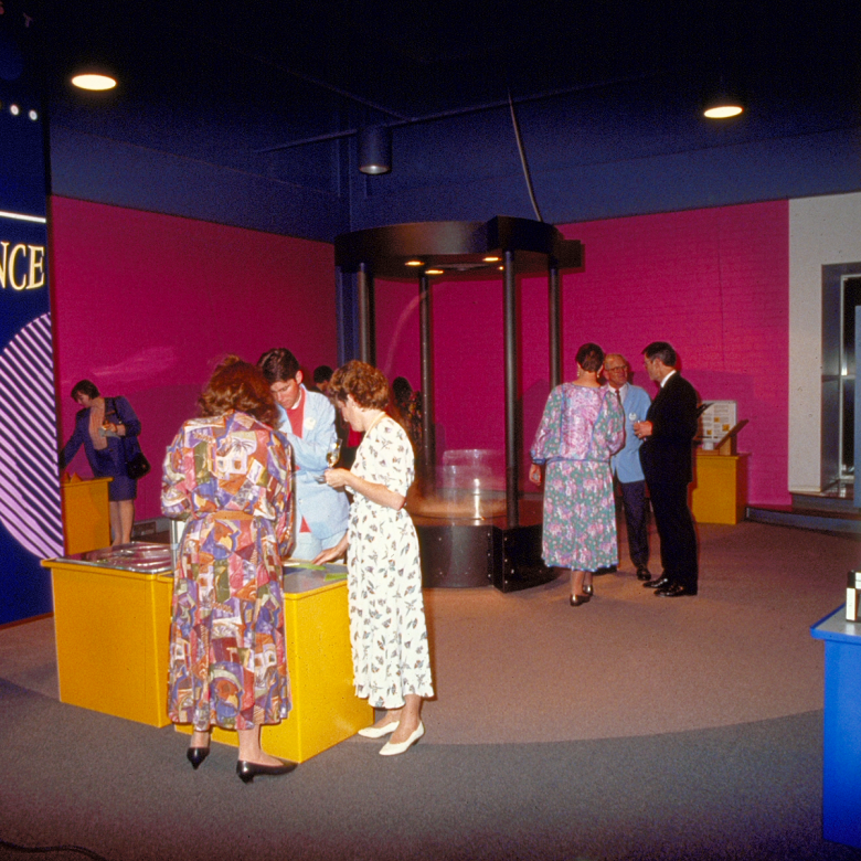 Adults in bright, patterned nineties fashion stand around colourful exhibits. A banner behind them reads 'FASCINATING SCIENCE'