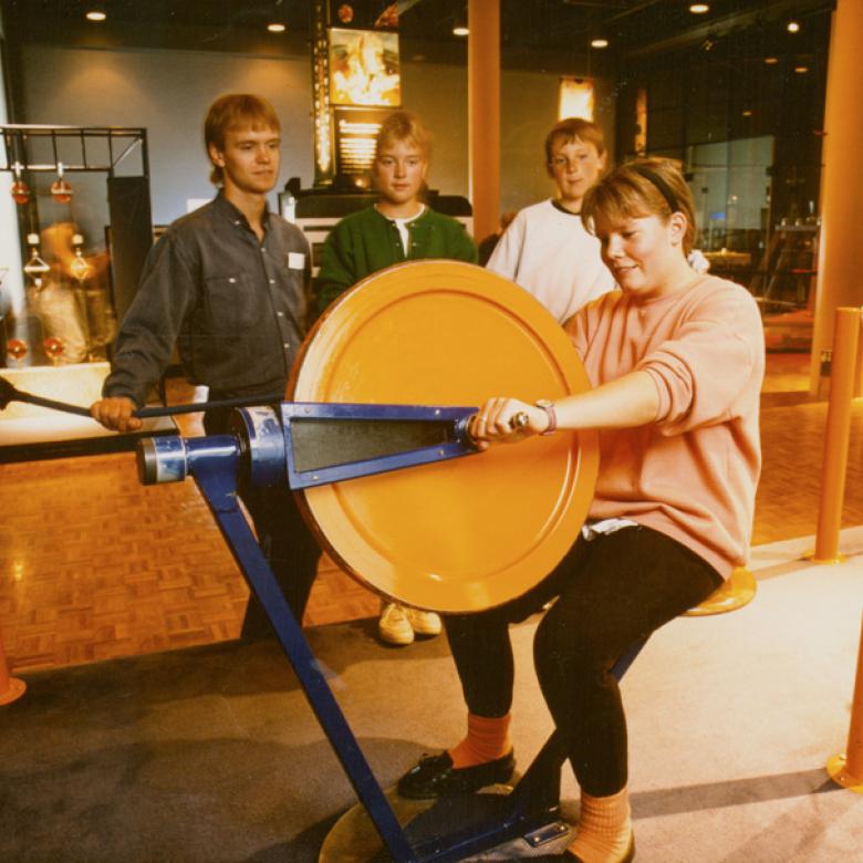 Three bystanders watch a woman use an exhibit by sitting on a chair connected to a huge orange gyroscope.