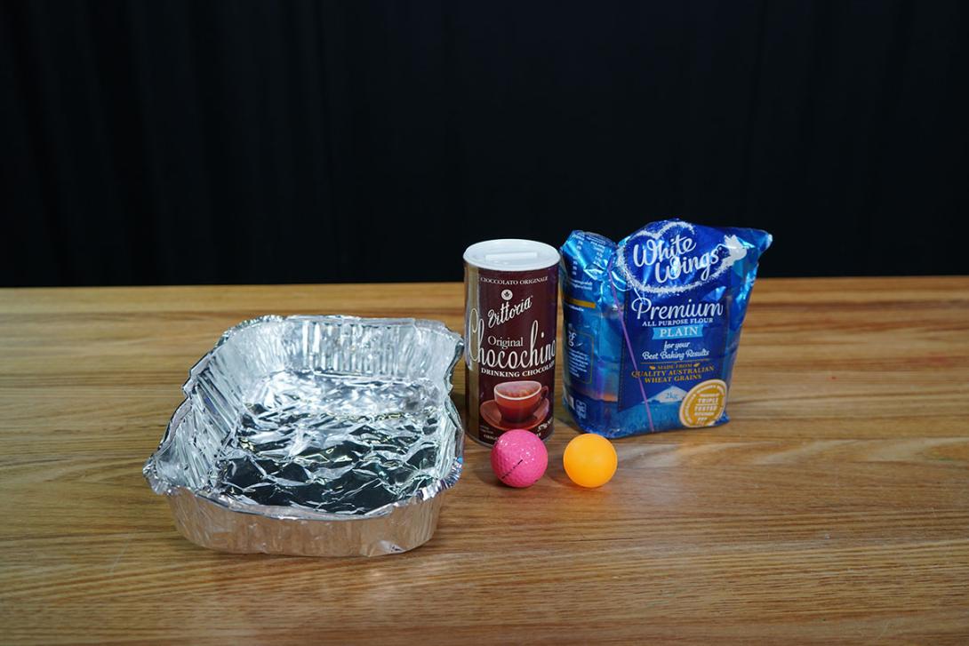 A disposable aluminium tray, a bag of flour, a tube of cocoa powder, a table tennis ball and a golf ball all sitting on a wooden table.