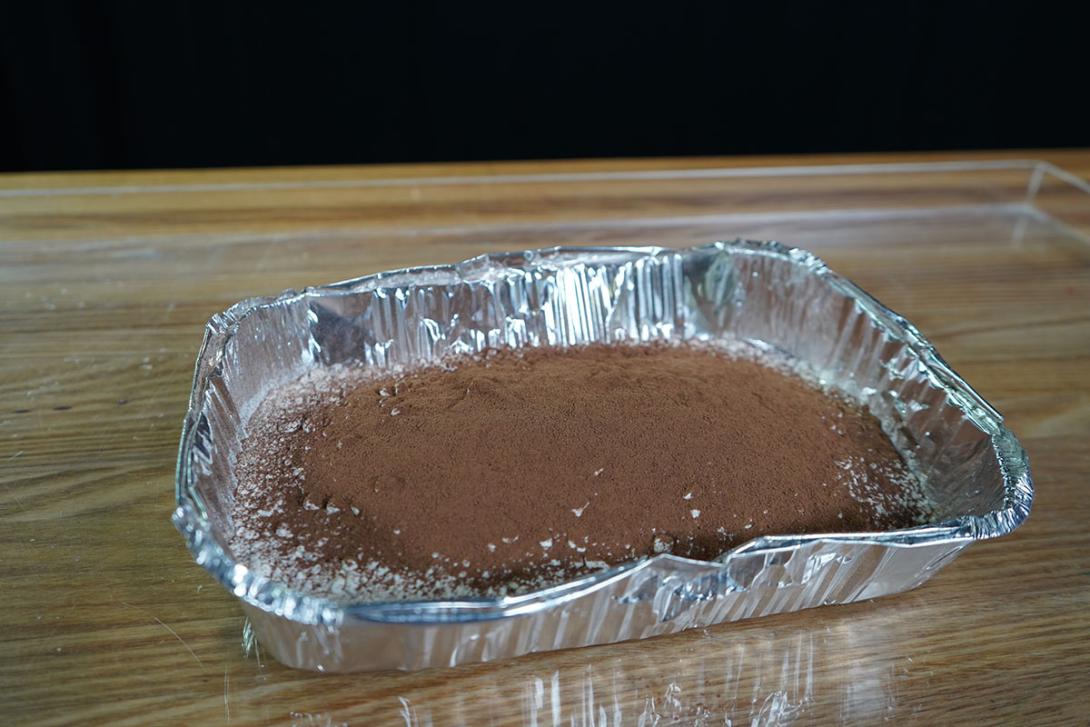 A disposable aluminium tray filled with flour several centimetres thick. The flour is covered by a light dusting of brown cocoa powder. 