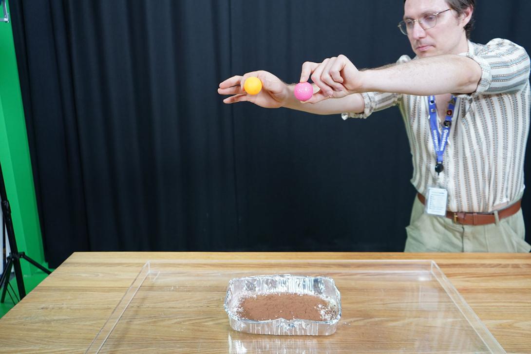 Hands holding an orange table tennis ball and a pink golf ball about a meter above a disposable aluminium tray filled with flour covered by a light dusting of brown cocoa powder. 